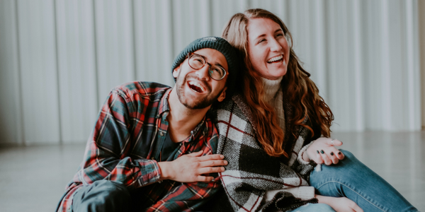 guy and girl in their 20's sitting in a barn and laughing, wearing a beanie and glasses, flannel and jeans.